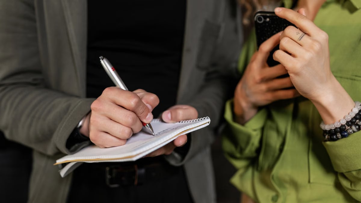 Two hands, one writing in a notebook, the other holding a smartphone, captured close-up.